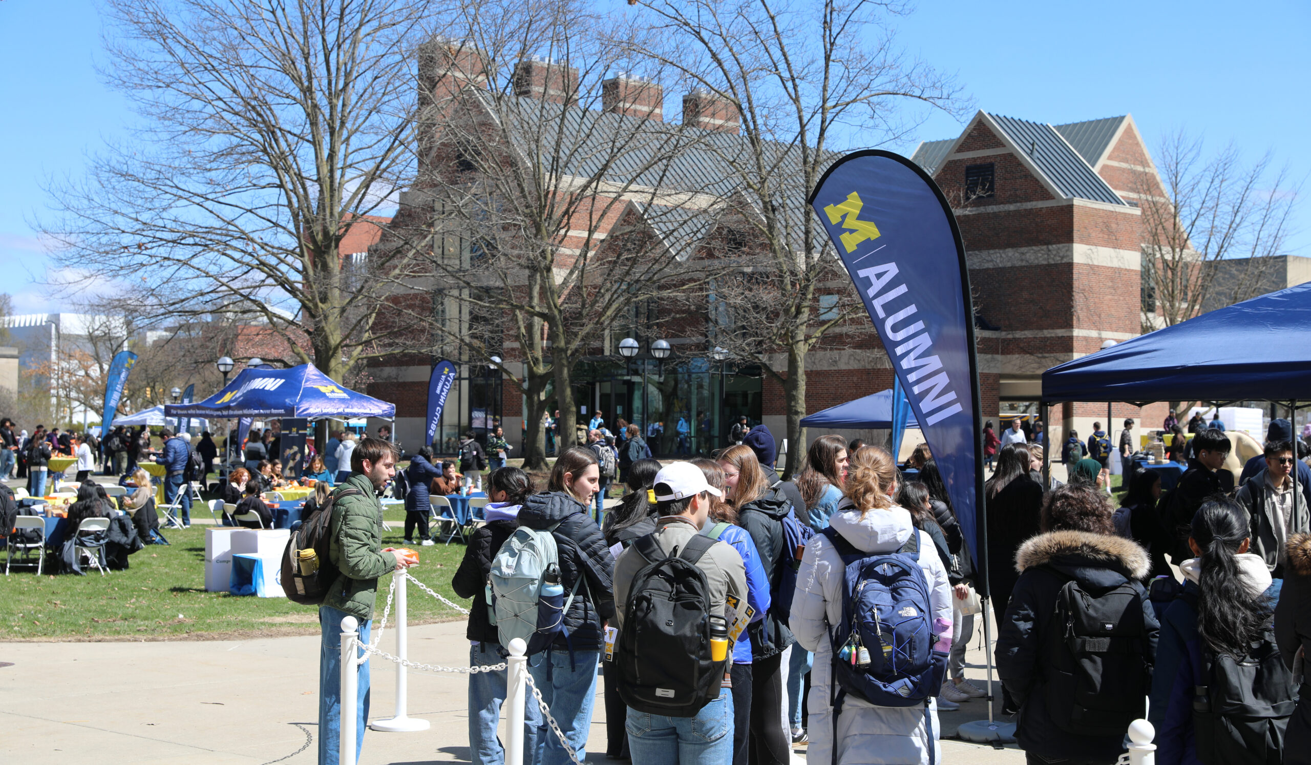 Students wait in various lines and gather in groups on Ingalls Mall. Many blue pop-up tents can be seen and the Alumni Center is in the background.